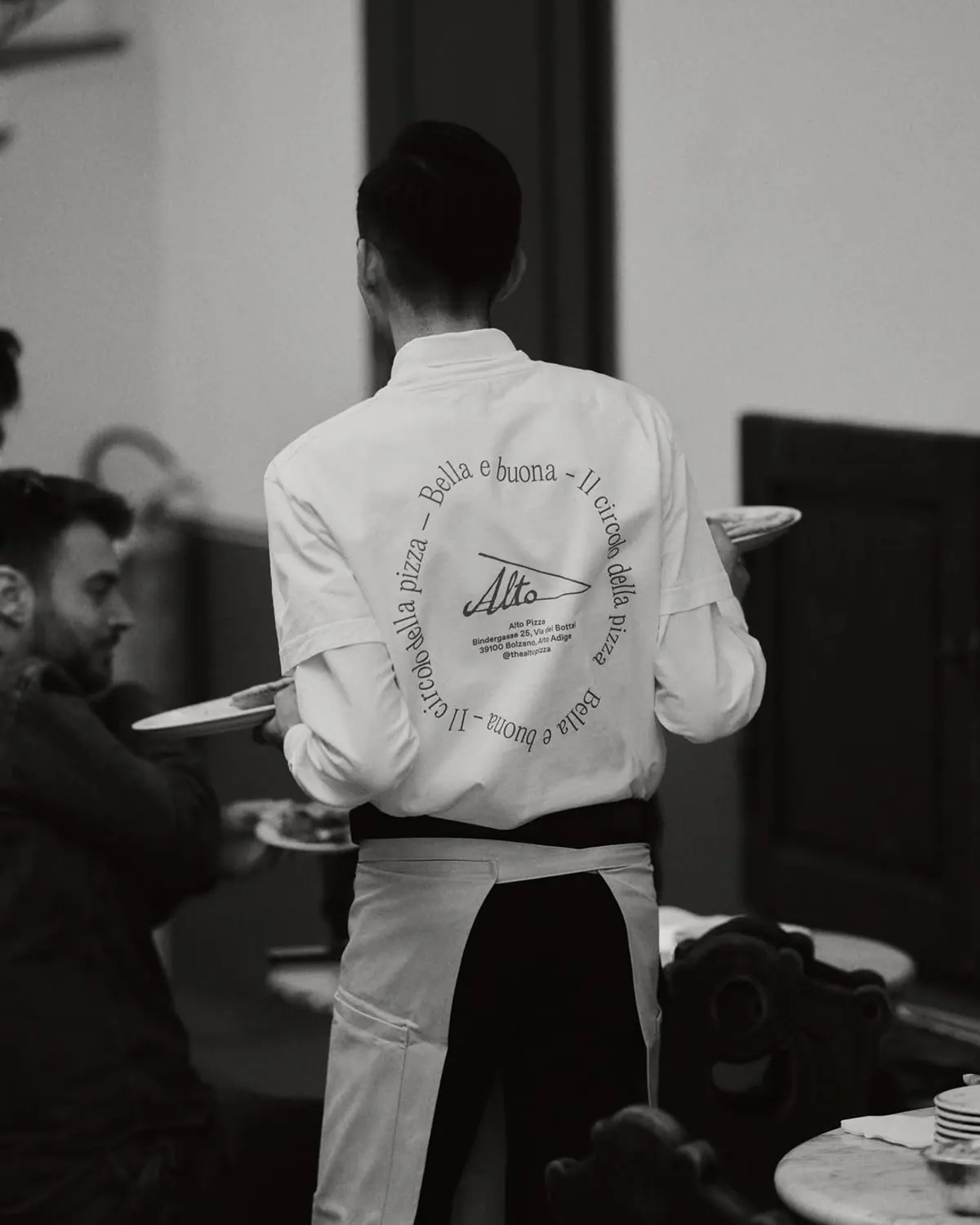 Restaurant server at Alto in a white chef-style coat with the Alto print on the back, carrying plates