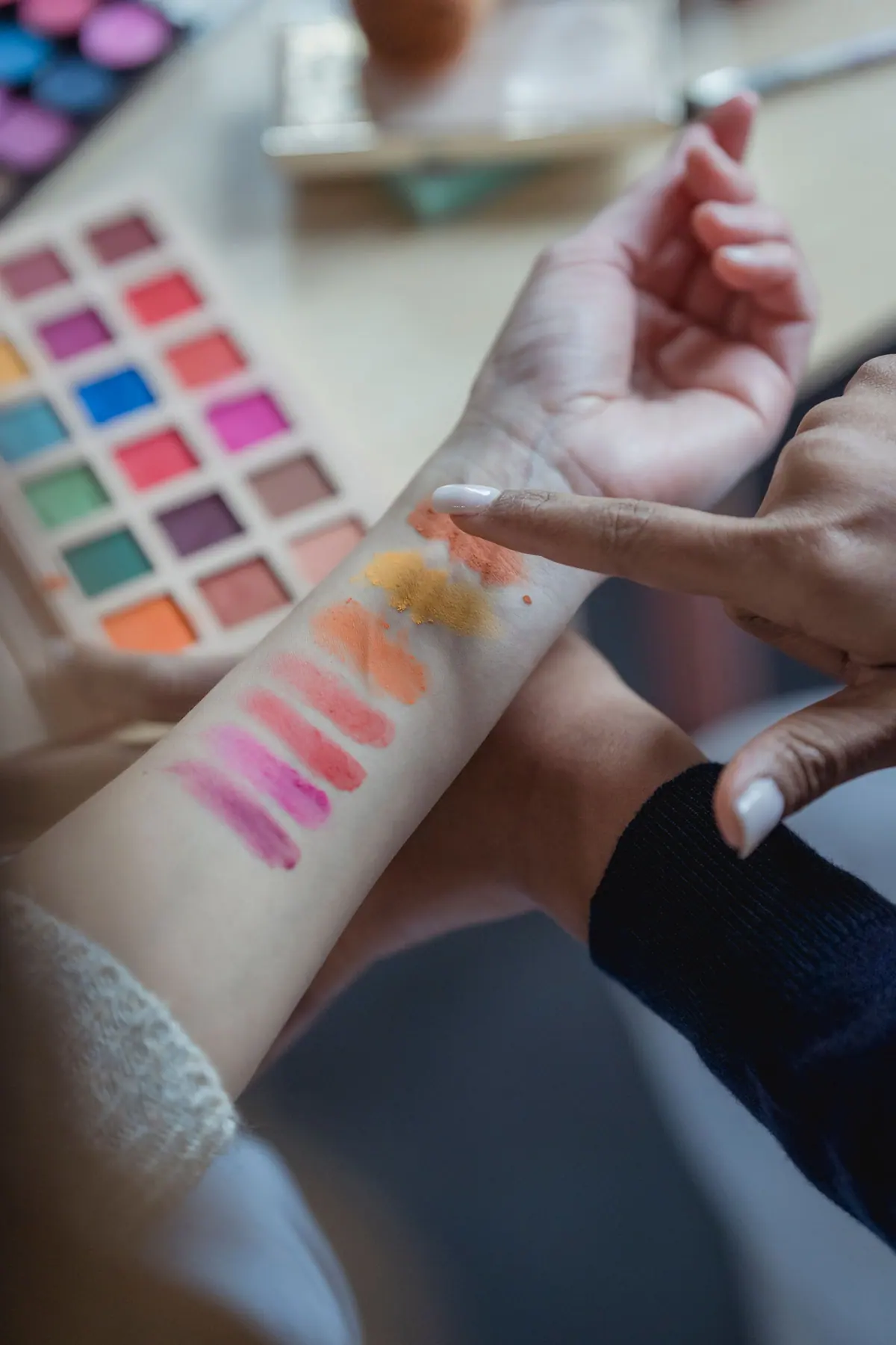 High angle of crop anonymous female visagiste applying various colorful eyeshadows on arm of client before doing makeup. Hands only: advisor swatches pigments on the client's forearm beside an eyeshadow palette. 4000x6000
