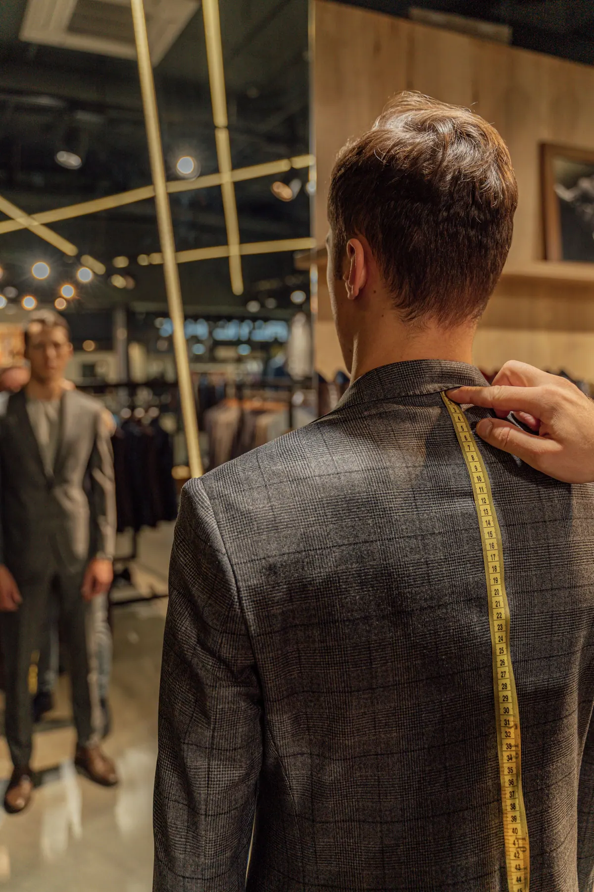 A man getting a checked blazer fitted; tailor's hand runs a tape measure across the client's back, seen from behind, reflected in a boutique mirror (4000x6000)