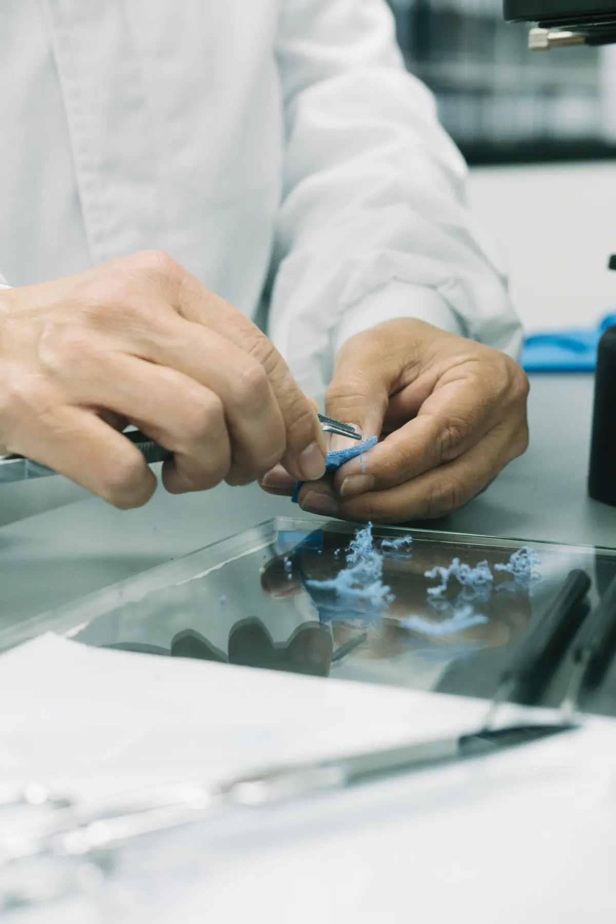 Hands in lab coats inspecting a blue fabric sample with tweezers over a glass surface