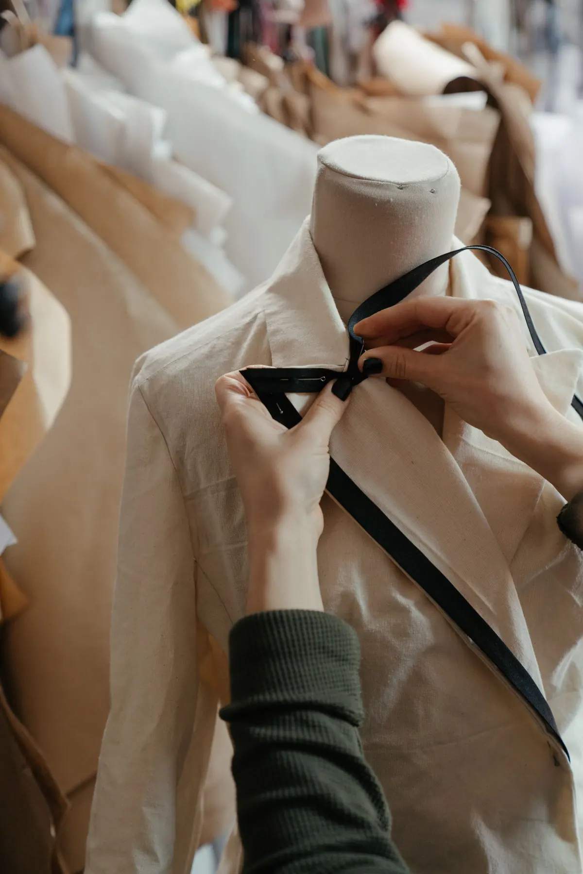Close-up of a tailor's hands adjusting a garment (white shirt with a black ribbon tie) on a dress form in a studio, with rows of forms behind (2333x3500)