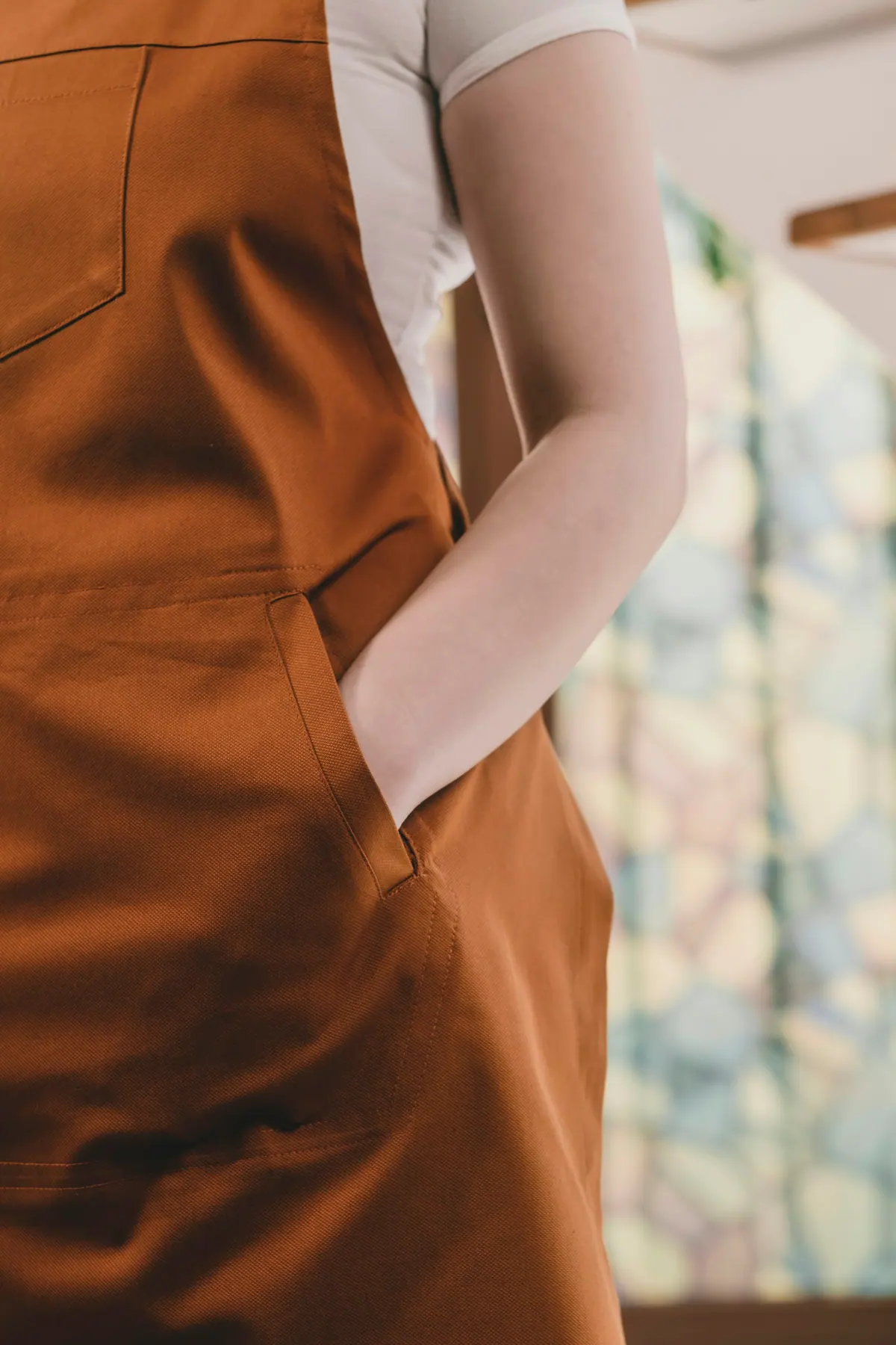 Rust cotton bib apron over a white shirt, hands in the pockets, waist-level crop